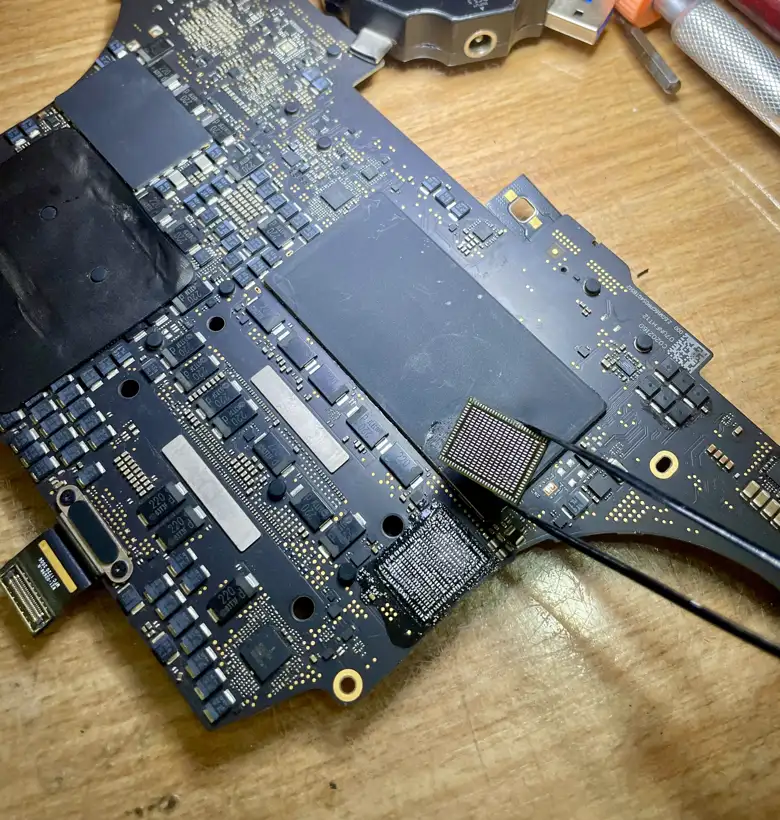 A MacBook Pro logic board on the bench with a BGA chip being lifted clear with tweezers, exposing the pad array.