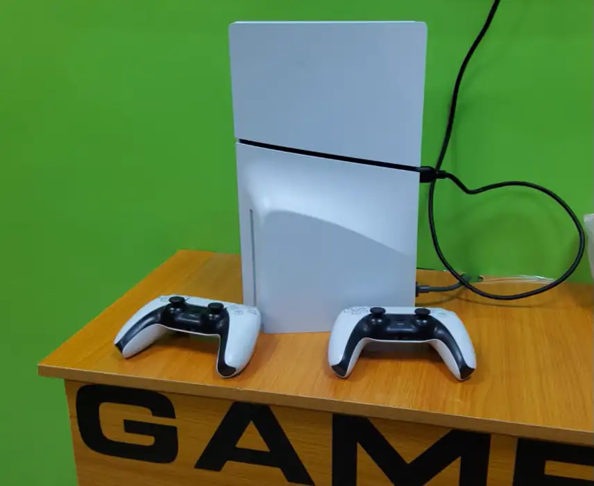 A white PlayStation 5 Slim standing on the Game Zone counter with two DualSense controllers.